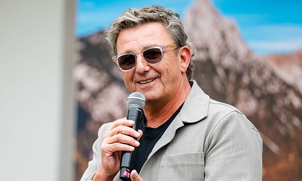 Mann mit Sonnenbrille und Mikrofon spricht auf einer Bühne, im Hintergrund unscharfe Berglandschaft.