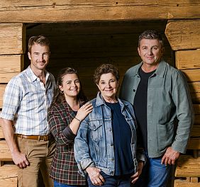 Eine Gruppe von vier Menschen steht vor einer hölzernen Wand. Sie lächeln und haben ihre Arme umeinander gelegt, in einer fröhlichen und herzlichen Pose.