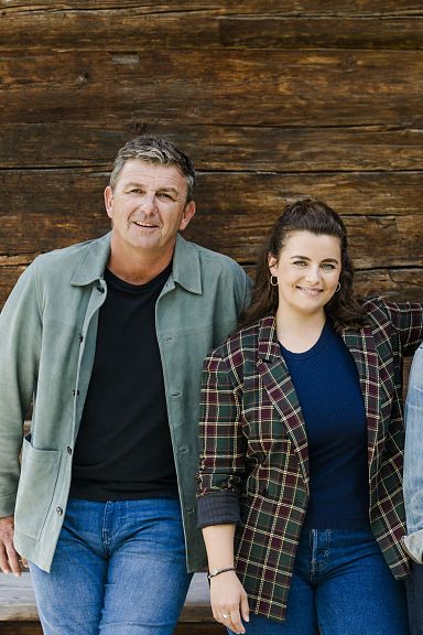 Vier Personen lehnen entspannt an einer Holzwand, kleiden sich in casualer Kleidung und lächeln in die Kamera. Die Atmosphäre ist freundlich und vertraut.