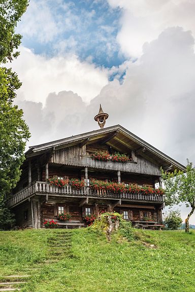 Holzhütte in Berglandschaft mit blauem Himmel, großen Bäumen und grüner Wiese. Traditionelle Architektur mit Blumen geschmücktem Balkon.