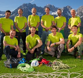 A group of people in bright green shirts pose with climbing gear outdoors, with a scenic mountain backdrop under a clear blue sky.