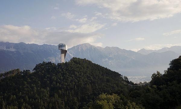 Turm auf einem bewaldeten Hügel vor einer Bergkulisse bei bewölktem Himmel. Im Hintergrund sind hohe Gipfel und ein Tal zu erkennen.