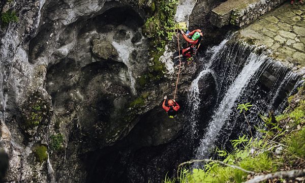 Two people in red gear rappel down a rocky waterfall surrounded by lush greenery near the Wilder Kaiser mountains, highlighting adventure and nature.