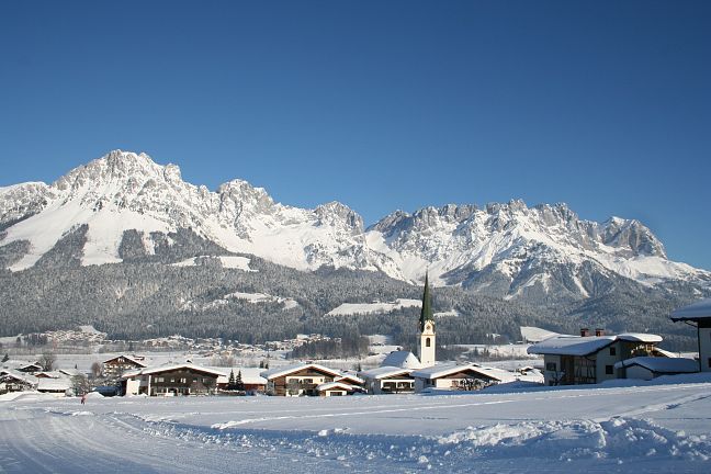 Verschneite Berglandschaft mit markantem Gipfel, darunter ein malerisches Dorf mit einer Kirche und mehreren Häusern, im Vordergrund eine Schneefläche.