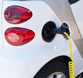 Ein weißes Elektroauto wird mit einem gelben Ladekabel an eine Ladestation angeschlossen. Im Hintergrund sind Rücklichter zu sehen. Nahaufnahme.