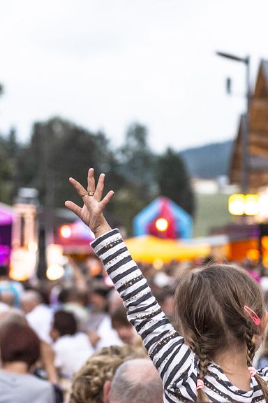 Outdoor concert scene with a large crowd and a young girl with raised arms enjoying the music. Colorful stage lights and tents add to the festive atmosphere.