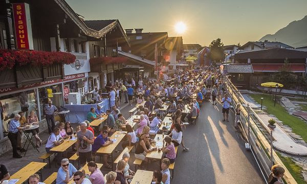 Menschen sitzen an langen Bierbänken im Freien, genießen Getränke und Essen bei Sonnenuntergang in einem lebhaften, traditionell gestalteten Dorf.