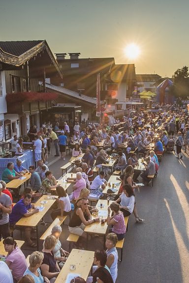 Menschen sitzen an langen Bierbänken im Freien, genießen Getränke und Essen bei Sonnenuntergang in einem lebhaften, traditionell gestalteten Dorf.
