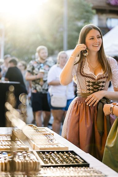 Zwei Frauen in traditionellen Dirndln betrachten ausgestellte Schmuckstücke auf einem Markt bei sonnigem Wetter, umgeben von anderen Besuchern.
