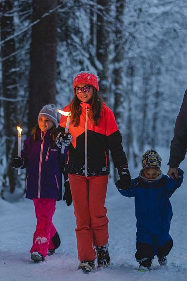 Familie spaziert im Winterwald bei Dämmerung, hält Fackeln und lächelt. Sie tragen warme Kleidung und genießen die verschneite Landschaft.