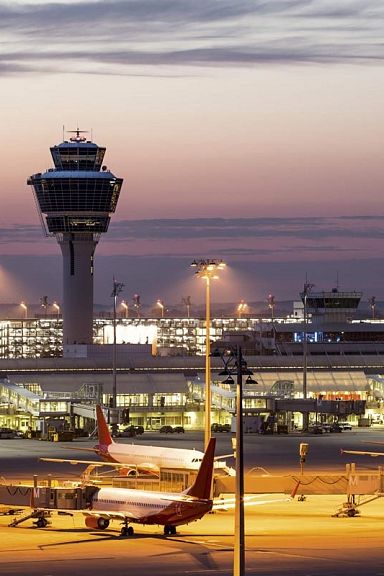 Ein Flughafen bei Dämmerung mit beleuchteten Rollfeldern, geparkten Flugzeugen und einem markanten Kontrollturm. Der Himmel zeigt Farbtöne von Orange und Lila.