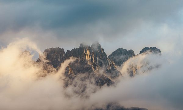 Misty mountain peaks of the Wilder Kaiser emerge through clouds, bathed in warm light, offering a dramatic and serene alpine scene.