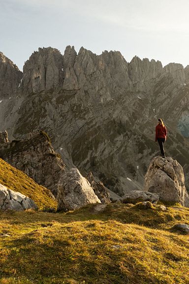 Eine Person steht auf einem Felsen mit Blick auf ein majestätisches Bergpanorama bei Sonnenuntergang. Die Berge sind karg und dramatisch geformt.