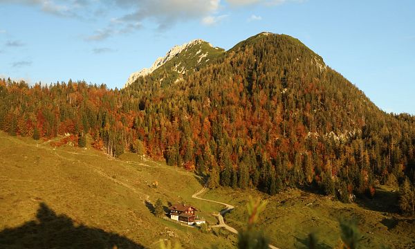 Herbstliche Berglandschaft mit bunten Bäumen, einem kleinen Haus im Tal und einem blauen Himmel. Die Sonne wirft warme Schatten auf die Szenerie.