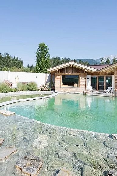 Natur-Schwimmteich mit klarer, grüner Wasserfarbe, umgeben von Natursteinen und hölzernen Gebäuden im Alpenstil, im Hintergrund Wald und Berge.