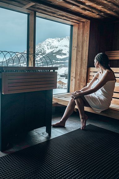 Frau sitzt in einer Sauna mit Holzinterieur und blickt durch ein großes Fenster auf schneebedeckte Berge und ein Tal in winterlicher Landschaft.