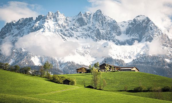 Eine malerische Berglandschaft mit schneebedeckten Gipfeln, grünen Wiesen und traditionellen Berghütten unter einem teils bewölkten Himmel in Tirol.