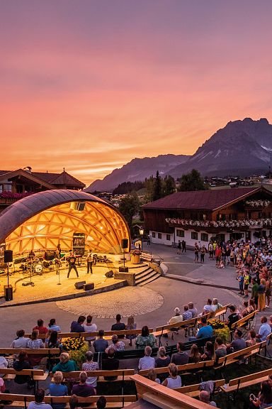 Ein Open-Air-Konzert in einem Bergdorf bei Sonnenuntergang. Zuschauer sitzen vor einer beleuchteten Bühne, umgeben von traditionellen Gebäuden und Bergen im Hintergrund.