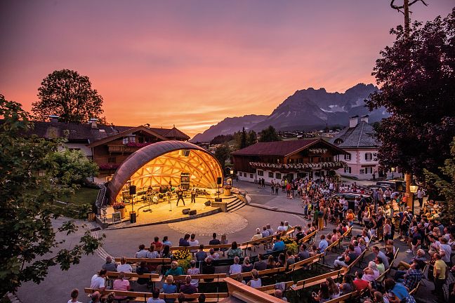Ein Open-Air-Konzert in einem Bergdorf bei Sonnenuntergang. Zuschauer sitzen vor einer beleuchteten Bühne, umgeben von traditionellen Gebäuden und Bergen im Hintergrund.
