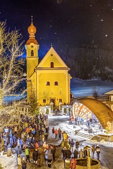 Winterlicher Kirchplatz mit beleuchteter Kirche und Menschenmenge bei Nacht, Schnee bedeckt den Boden, ein großer Baum neben der Kirche.