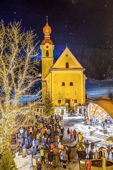 Winterlicher Kirchplatz mit beleuchteter Kirche und Menschenmenge bei Nacht, Schnee bedeckt den Boden, ein großer Baum neben der Kirche.