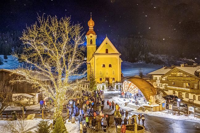 Winterlicher Kirchplatz mit beleuchteter Kirche und Menschenmenge bei Nacht, Schnee bedeckt den Boden, ein großer Baum neben der Kirche.