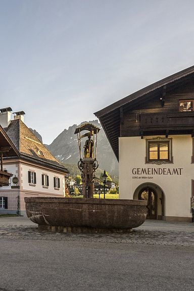 Ein malerischer Dorfplatz mit traditionellen Tiroler Holzhäusern und einem Brunnen in der Mitte, umgeben von Bergen und blauem Himmel im Hintergrund.