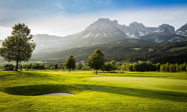 Grüner Golfplatz mit Sandbunker, umgeben von Bäumen. Im Hintergrund erhebt sich majestätisch ein beeindruckendes Bergmassiv unter blauem Himmel.