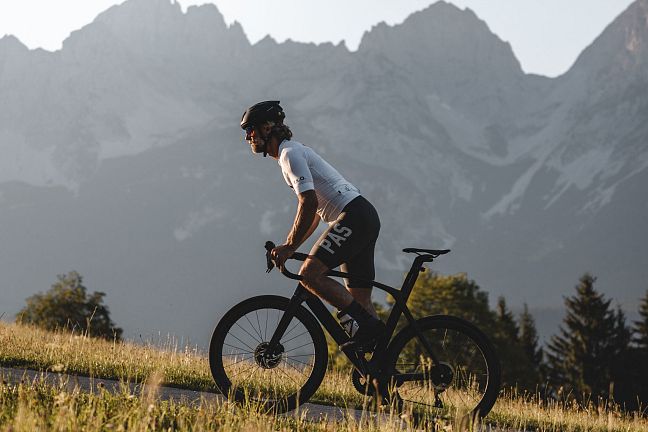 Ein Radfahrer in sportlicher Kleidung fährt über eine grüne Wiese. Im Hintergrund erheben sich eindrucksvolle Berggipfel gegen den klaren Himmel.