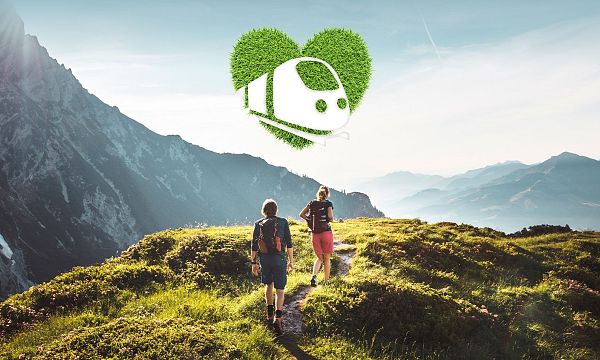 Zwei Menschen wandern auf einem grünen Bergpfad mit malerischer Berglandschaft im Hintergrund. Über ihnen schwebt ein Zug-Symbol in einem herzförmigen Blattmuster.