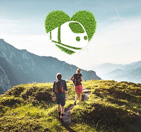 Two people hike on a green mountain trail with the Wilder Kaiser mountains in the background, under a blue sky with a heart-shaped eco symbol overhead.