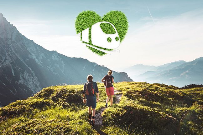 Two people hike on a green mountain trail with the Wilder Kaiser mountains in the background, under a blue sky with a heart-shaped eco symbol overhead.