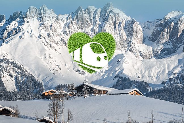 Verschneites Bergmassiv des Wilden Kaisers mit traditionellen Holzhütten im Vordergrund. Ein Herz-Symbol aus Gras mit einem Zug-Icon ist überlagert.