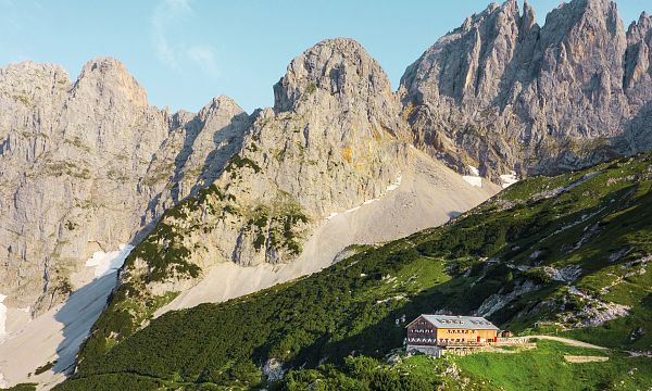 Berglandschaft mit markanten Felsformationen und einer Berghütte im Vordergrund. Der Himmel ist klar und blau, umgeben von grünen Almen und steilen Hängen.