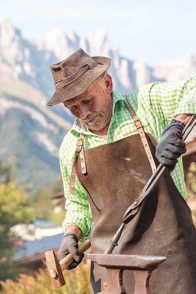 Ein älterer Mann in traditioneller Arbeitskleidung arbeitet draußen an einem Amboss, vor einer malerischen Berglandschaft mit Holzhäusern.