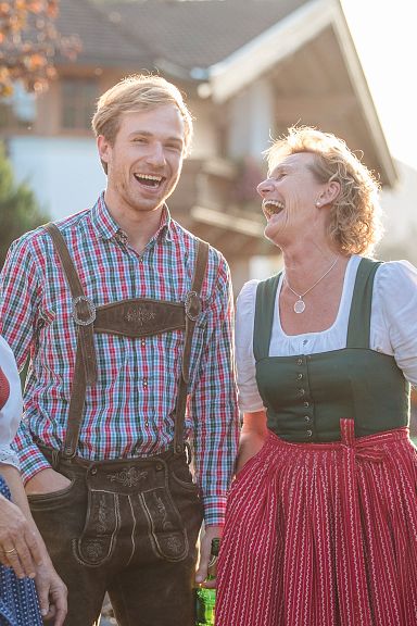 Drei Personen in traditioneller österreichischer Tracht lachen und unterhalten sich im Freien vor einem Haus. Die Sonne scheint, und andere Personen sind im Hintergrund.