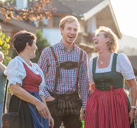 Drei Personen in traditioneller österreichischer Tracht lachen und unterhalten sich im Freien vor einem Haus. Die Sonne scheint, und andere Personen sind im Hintergrund.