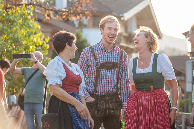 Three people in traditional attire laughing outdoors with a blurred background of trees and a house.