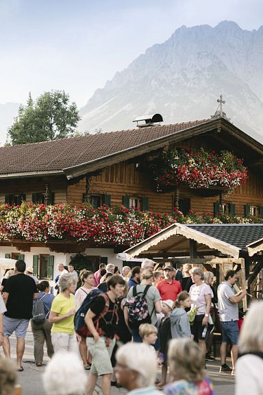 Menschenmenge auf einem belebten Fest vor einem traditionellen Holzhaus mit Blumen, im Hintergrund ein beeindruckendes Bergpanorama.