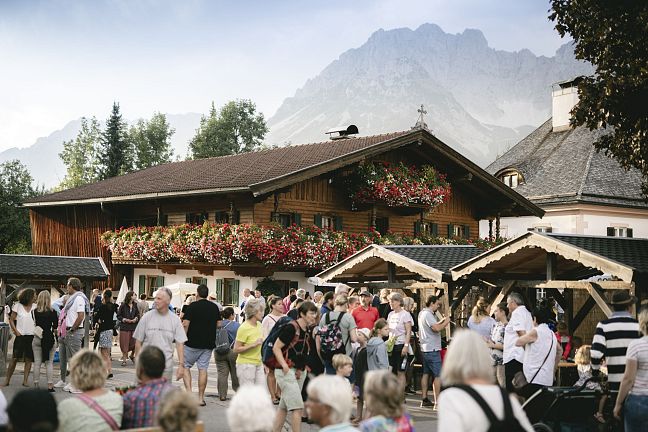 Menschenmenge auf einem belebten Fest vor einem traditionellen Holzhaus mit Blumen, im Hintergrund ein beeindruckendes Bergpanorama.