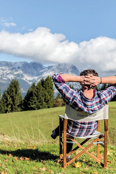 Person sitzt entspannt auf einem Stuhl auf einer Wiese, blickt auf ein beeindruckendes Bergpanorama mit bewaldeten Hängen und blauem Himmel im Hintergrund.