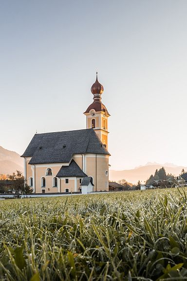 Eine malerische Kirche mit Zwiebelturm steht auf einer grünen Wiese, umgeben von Hügeln und Bergen unter klarem Himmel.