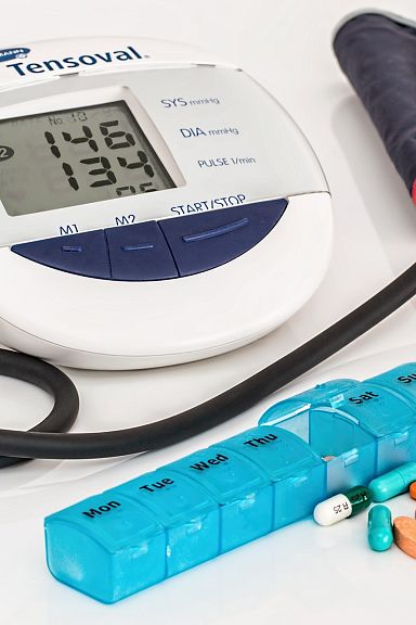 Blood pressure monitor with cuff, daily pill organizer, and assortment of colorful tablets and capsules on a white surface.