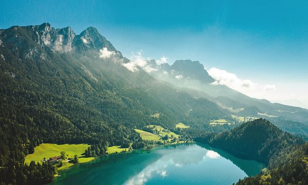 Ein klarer, türkisblauer See umgeben von grünen Wäldern und majestätischen Bergen unter klarem, blauem Himmel. Ein idyllisches Landschaftspanorama.
