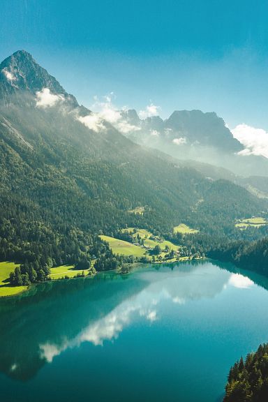 Ein klarer, türkisblauer See umgeben von grünen Wäldern und majestätischen Bergen unter klarem, blauem Himmel. Ein idyllisches Landschaftspanorama.