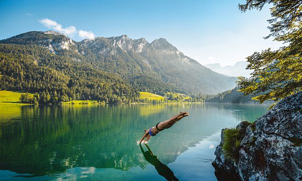Person taucht in klaren Bergsee mit Wald und Bergen im Hintergrund. Sonniges Wetter, ruhige Atmosphäre, grüne Umgebung und malerische Landschaft.