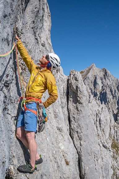 A climber in a yellow jacket navigates a steep rock face in the Wilder Kaiser region, with jagged mountain peaks visible in the background.