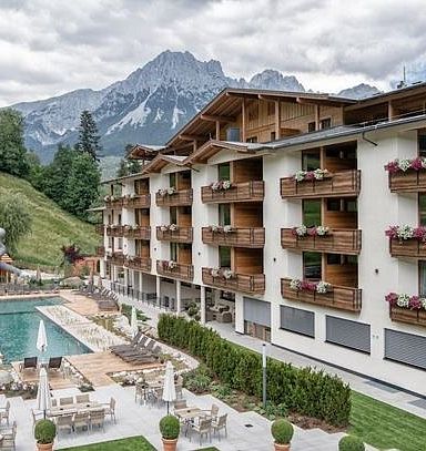 A hotel with wooden balconies overlooks a pool and patio area, with the Wilder Kaiser mountains in the background under a cloudy sky.
