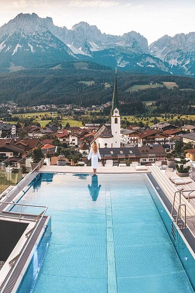 A woman in a white robe stands by a rooftop pool, overlooking a historic town with a church, set against the backdrop of lush green mountains, Wilder Kaiser.