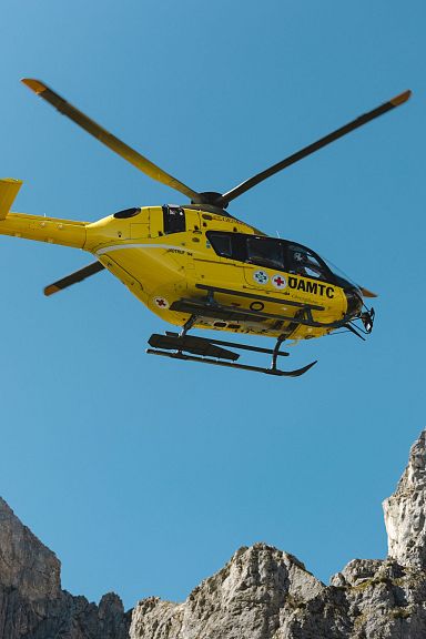 Gelber Rettungshubschrauber fliegt vor blauem Himmel über felsigem Bergmassiv in den Alpen. Österreichische Flagge am Heck des Helikopters sichtbar.